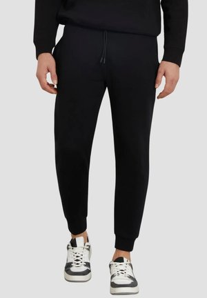 Hombre con pantalones jogger negros y zapatillas negras y blancas, de pie frente a un fondo liso y claro.