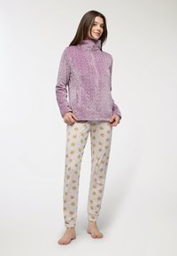 Pullover in pile di tessuto viola con colletto alto e tasche frontali, abbinato a pantaloni crema con un motivo a ripetizione di facce di cartone animato.
