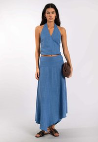 Mujer con top corto halter azul y falda maxi asimétrica a juego, sosteniendo un clutch marrón texturizado, usando sandalias negras con tiras.