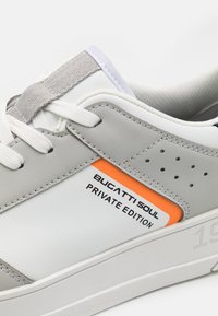 Sneaker in pelle bianca e grigia con dettagli forati, etichetta arancione con la scritta "BUGATTI SOUL PRIVATE EDITION" e suola testurizzata.