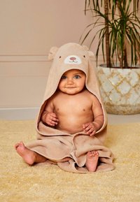 Teddy bear hooded towel in beige, soft terrycloth material. Towel drapes around a seated child, with facial detailing on the hood.