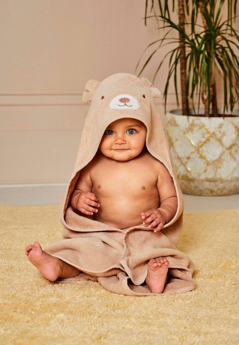Teddy bear hooded towel in beige, soft terrycloth material. Towel drapes around a seated child, with facial detailing on the hood.