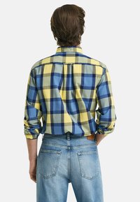 Herren karierter Kurzarmhemd in Gelb, Blau und Grün, mit hochgekrempelten Ärmeln und einem Rückenfalten. Getragen mit hellblauen Jeans.