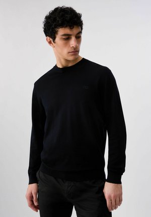 Uomo con capelli ricci che indossa un maglione nero a maniche lunghe semplice e pantaloni scuri, che guarda in basso verso sinistra su uno sfondo neutro.