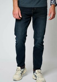 Tmavě modré džíny s úzkým střihem, vybavené jemnými švy a klasickým pětiprvkovým designem kapes. Doplněné bílými sportovními botami.