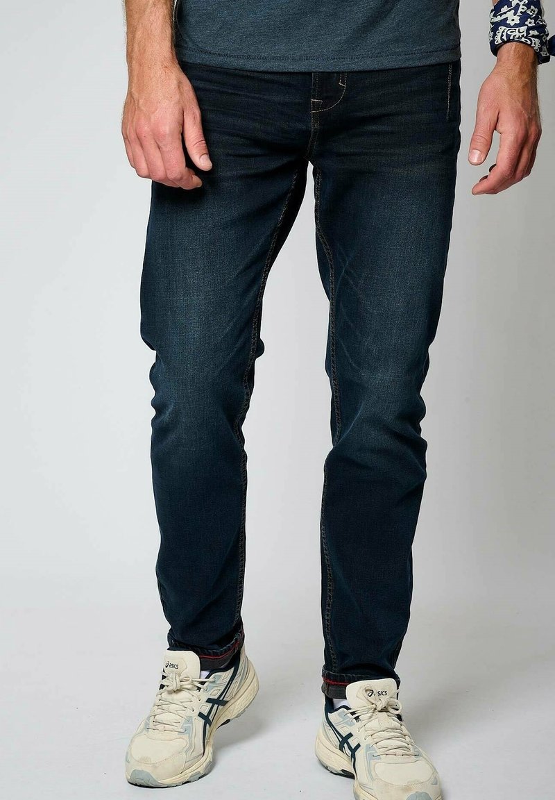 Tmavě modré džíny s úzkým střihem, vybavené jemnými švy a klasickým pětiprvkovým designem kapes. Doplněné bílými sportovními botami.