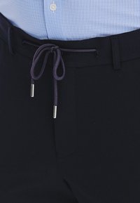 Granatowe spodnie z elastycznym sznureczkiem, wykonane z teksturowanej tkaniny, z fioletowym sznurkiem i metalowymi końcówkami. Pokazane z jasnoniebieską koszulą w kratę.