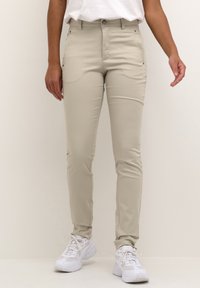 Bež hlače slim fit izrađene od rastezljive tkanine, s prednjim džepovima i zatvaranjem na gumb, u paru s bijelim sportskim tenisicama.