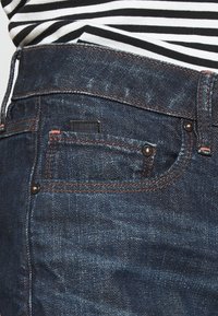 Tmavě modré džíny z denimu mají oranžové prošívání, klasický design s pěti kapsami a malou černou nášivku na zadní kapse. Textura je mírně opotřebovaná.