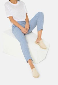 Béžové espadrilky s jemnou texturou, světle modré vzorované kalhoty a průhledný bílý tričko, sedící na bílém geometrickém bloku.
