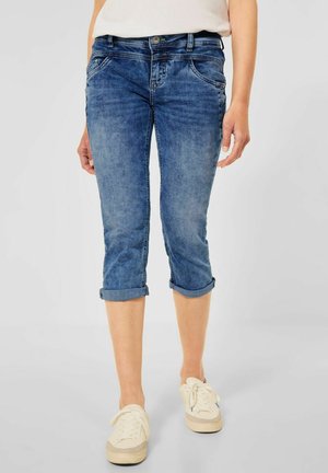 Person, die hochgekrempelte blaue Denim-Caprihosen und weiße Turnschuhe trägt, steht vor einem schlichten hellen Hintergrund.