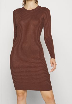 Femme portant une robe marron en maille côtelée, ajustée, à manches longues, tombant au-dessus des genoux, sur un fond clair uni.