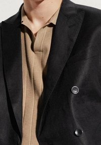 Dwurzędowa czarna marynarka o gładkiej fakturze, zestawiona z brązową koszulą w prążki. Posiada wyraźne guziki i wyprofilowany kołnierz.