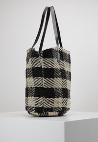 Schwarze und beige gewebte Tragetasche mit einem karierten Muster und zwei langen schwarzen Lederschulterriemen, die auf einem weißen Podest hängen.