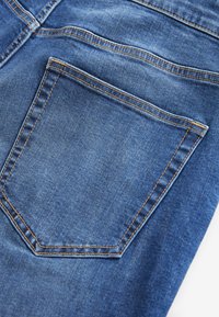 Jeans aus Denim mit einer Gesäßtasche in markanter dreieckiger Form, abgeschlossen mit hellbraunen Nähten auf mittlerem Blau.
