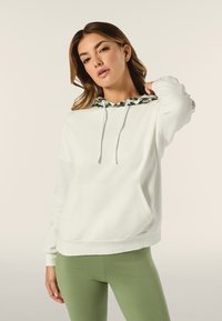 Felpa bianca con cappuccio a motivo verde e nero, cordini, tasca frontale e polsini a coste, abbinata a leggings verdi.