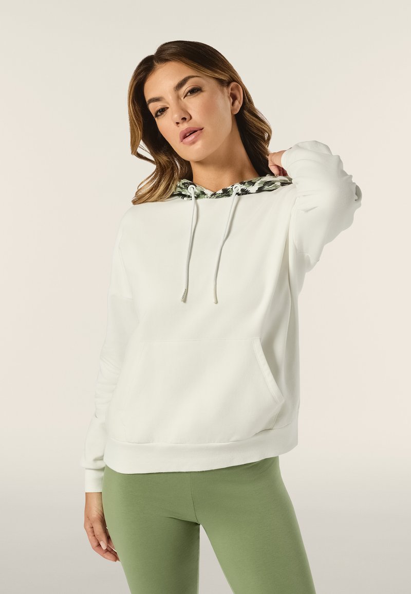 Felpa bianca con cappuccio a motivo verde e nero, cordini, tasca frontale e polsini a coste, abbinata a leggings verdi.