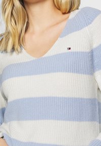 Maglione lavorato a maglia con scollo a V caratterizzato da strisce alternate di blu chiaro e bianco. Piccolo logo rosso, bianco e blu sul petto.
