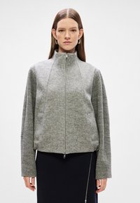 Γκρίζα ζακέτα με φερμουάρ και υψηλό γιακά, υφή υφάσματος και ραμμένες λεπτομέρειες στα μανίκια. Διαθέτει δύο πλευρικές τσέπες και κομμένη σιλουέτα.