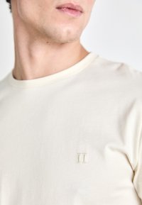 Kreminės spalvos trikampė rankovė t-shirt, pagaminta iš lygaus medvilnės, su mažu siuvinėtu "II" logotipu kairėje krūtinės pusėje. Išlenktas apykaklės iškirpimas.