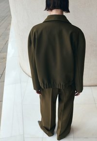 Henkilö seisoo selinpäin, yllään löysä oliivinvihreä takki ja siihen sopivat leveälahkeiset housut, laatoitetulla lattialla käyrän seinän lähellä.