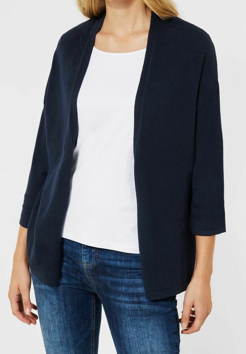 Laivastonsininen neuletakki pehmeästä kankaasta, jossa on kolme neljäsosahihnaa ja avoin etuosa. Yhdistetty valkoisen topin ja farkkujen kanssa. Sileä rakenne, rento istuvuus.