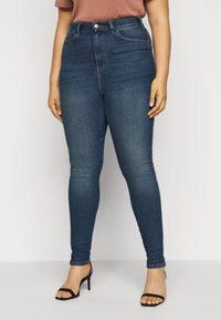 Person i mörkblå högmidjade skinnyjeans och svarta klacksandaler, stående mot en enkel ljus bakgrund.