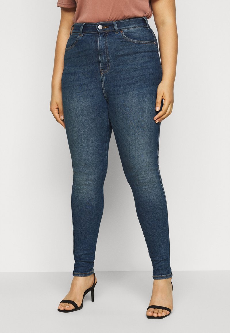 Person i mörkblå högmidjade skinnyjeans och svarta klacksandaler, stående mot en enkel ljus bakgrund.