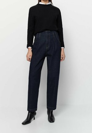 Femme portant un jean bleu foncé taille haute à jambes larges, un pull noir par-dessus une chemise blanche, et des bottines noires, debout sur fond blanc.