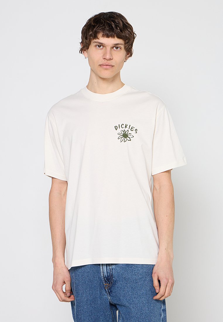 Junger Mann mit kurzem lockigem Haar, trägt ein weißes Dickies T-Shirt mit Blumenmotiv und blaue Jeans, steht vor einem schlichten Hintergrund.