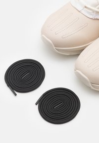 Des chaussures de sport beiges présentent un dessus texturé avec des rainures horizontales et des semelles en caoutchouc. Deux lacets ronds noirs avec embouts sont placés à proximité.