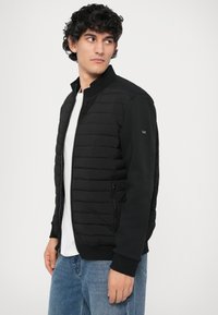 Casaco bomber preto com frente acolchoada, mangas em contraste, bolsos com fecho e punhos em canelado. Usado por cima de uma t-shirt branca e jeans azuis.
