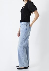 Femme portant un haut cache-cœur noir à manches bouffantes, un jean large taille haute bleu clair, et des ballerines noires pointues avec des détails en perles.