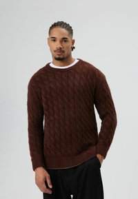 Bruine kabelgebreide trui van pluizig materiaal, met een ronde hals en lange mouwen, gecombineerd met een zwarte broek.