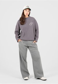 Un sweat à capuche gris en tricot avec un logo sur la poitrine, associé à un pantalon large gris clair et une casquette blanche avec des accents verts.