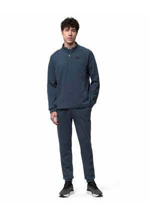Jeune homme portant un sweat-shirt bleu foncé à fermeture éclair quart de zip et un pantalon de survêtement assorti avec des baskets noires, debout sur fond blanc.