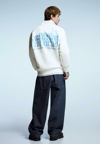 Cream sweatshirt with a high collar and blue graphic text on the back, paired with loose dark denim pants and black sneakers.