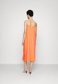 Oranžové midi šaty bez rukávů vyrobené z lehkého materiálu. Mají nízký zadní díl a boční rozparek, doplněné béžovými sandály na podpatku.
