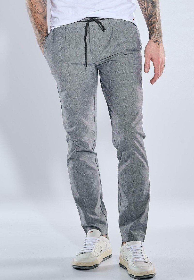 Graue, schmal zulaufende Hose aus einem gewebten Stoff, mit Seitentaschen und einem schwarzen Kordelzug. Tragen mit weißen Sneakern.