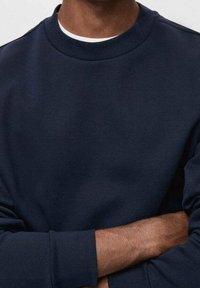 Sudadera de cuello redondo de color azul marino con mangas largas, tejido texturizado y escote liso, llevada con los brazos cruzados.