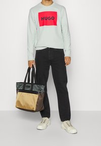 Felpa grigia chiara con un rettangolo rosso e il logo "HUGO" in nero. Jeans neri. Porta una borsa a tracolla bicolore in tonalità beige, marrone e verde.