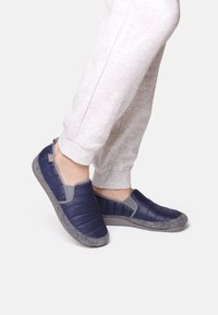 Marina slip-on tofflor med vadderad ovansidan i kviltat material och en grå filtliknande textur på sulan; slät yta med elastiska sidoflikar för enkel användning.