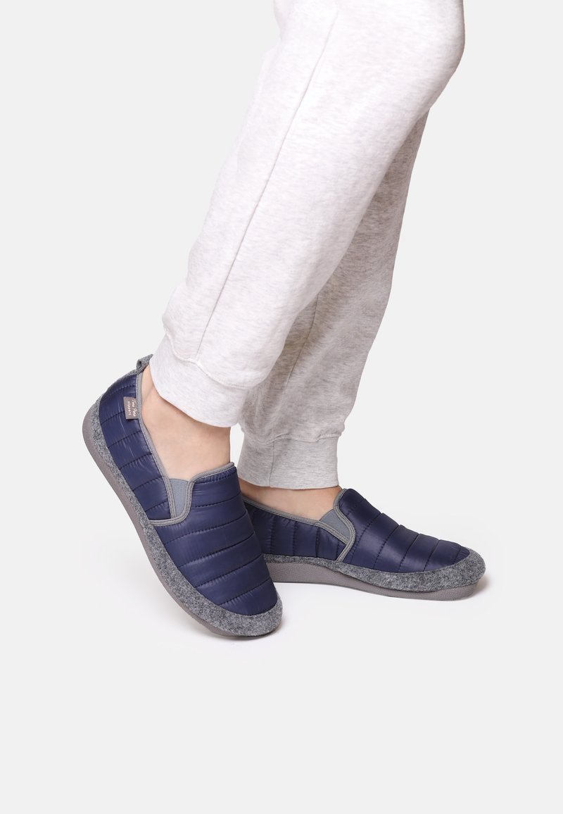 Marina slip-on tofflor med vadderad ovansidan i kviltat material och en grå filtliknande textur på sulan; slät yta med elastiska sidoflikar för enkel användning.