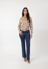 Beige gebreide cardigan met V-hals en gouden knopen, gecombineerd met blauwe rechte jeans. De cardigan heeft een gestructureerd patroon en voorzakken.