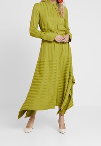 Robe verte à manches longues avec un col et une texture rayée. Présente un ourlet asymétrique et une taille cintrée. Portée avec des talons beiges.