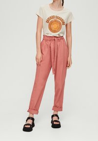 Camiseta de color beige claro con un gráfico de sol, combinada con pantalones de lino rosa de cintura alta con un cinturón y puños enrollados, llevados con sandalias negras.