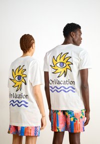 T-shirts de algodão brancas com um gráfico de um sol amarelo e um olho azul nas costas, apresentando o texto "Em Férias" e um design de onda azul abaixo.