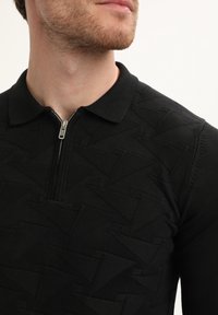 Vyras, vilkintis juodą mezginį polo marškinėlius su tekstūriniu zigzaginiu raštu ir pusiau užtrauktuku, rodantis apatinę veido dalį ir kaklą.