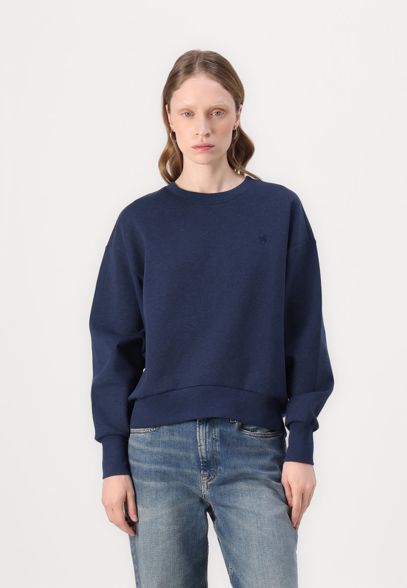 Marineblå genser med lange ermer, ribbestrikket kant og rund hals. Subtil logo på brystet. Kombinert med lyseblå jeans.