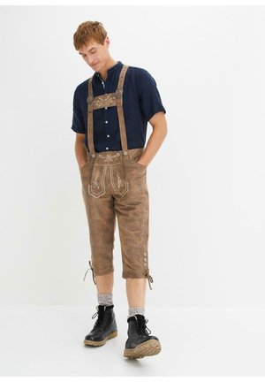 Lederhosen en cuir marron avec des détails brodés, des bretelles ajustables, deux poches et des ourlets roulés. Associé à une chemise sombre et des bottes noires.